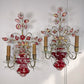 A pair of ornate antique Maison Bagues French wall sconces with red crystal glass vases, pink and red floral accents, gold-toned metal arms, and clear crystal prisms, shown against a plain white wall.