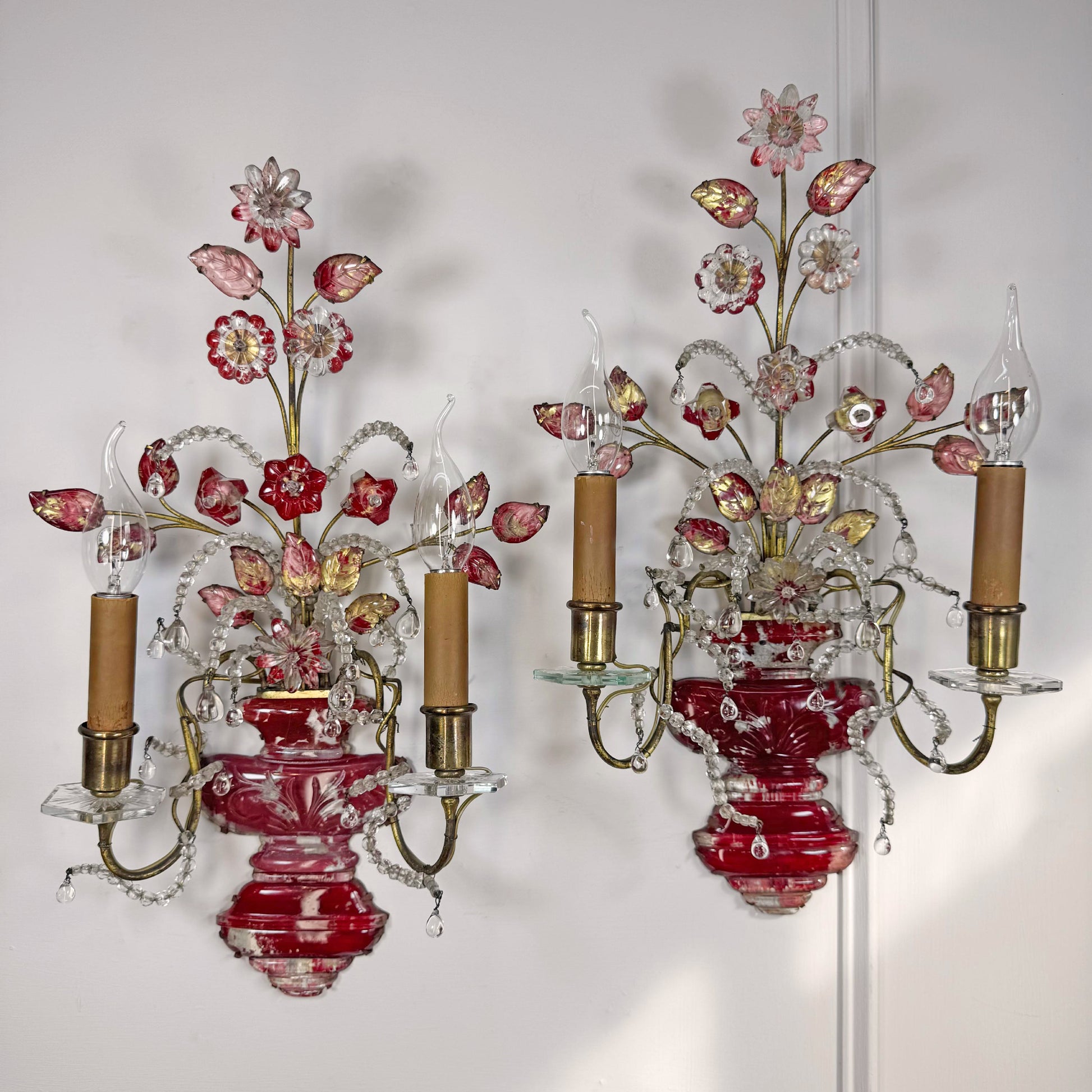 A pair of ornate antique Maison Bagues French wall sconces with red crystal glass vases, pink and red floral accents, gold-toned metal arms, and clear crystal prisms, shown against a plain white wall.