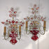 A pair of ornate antique Maison Bagues French wall sconces with red crystal glass vases, pink and red floral accents, gold-toned metal arms, and clear crystal prisms, shown against a plain white wall.