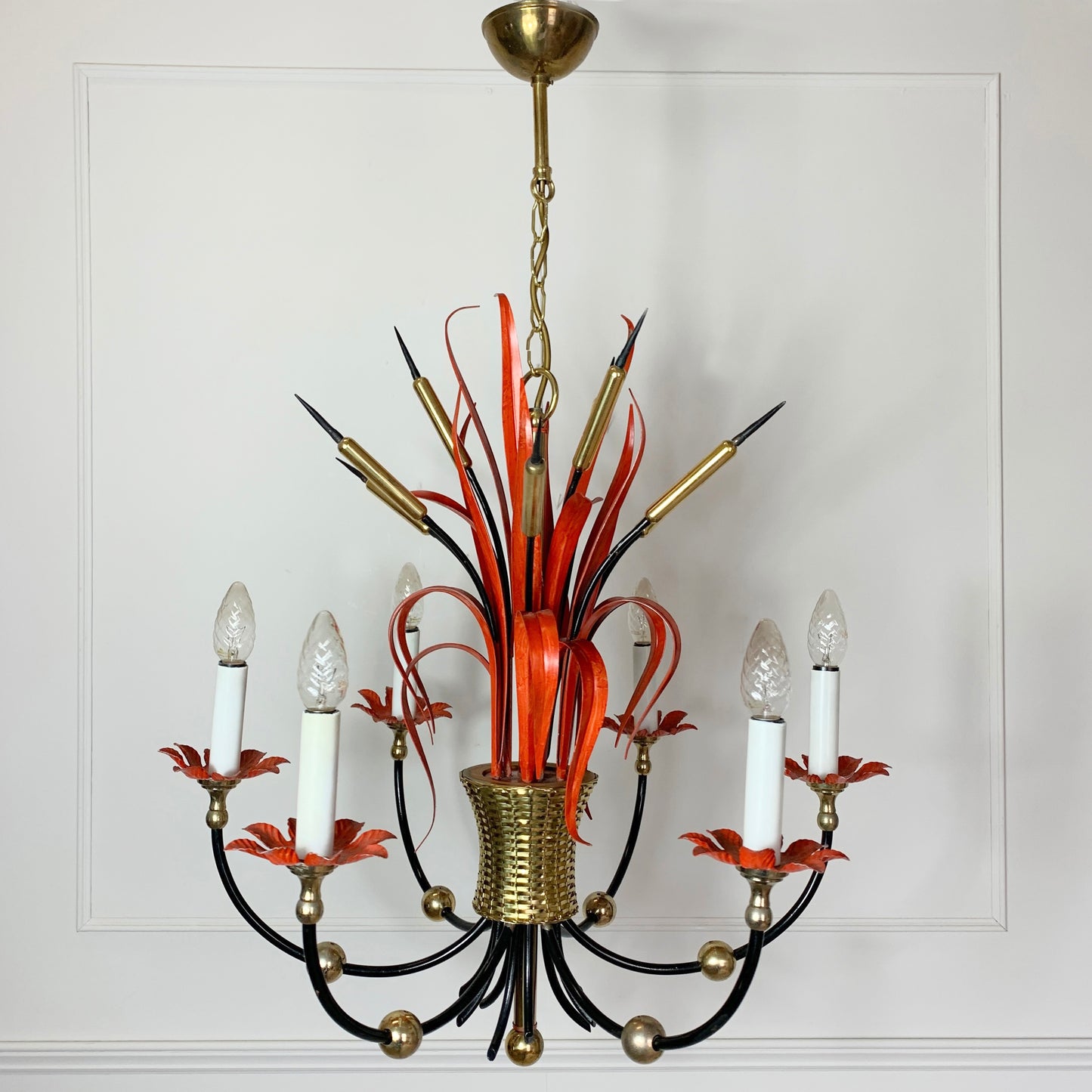 A vintage Maison Bagues Att Red Bulrush Chandelier with a gold-toned brass canopy and central part, black curved arms, orange-red ribbon-like accents, and clear glass candle-style bulbs, shown hanging against a white paneled wall.