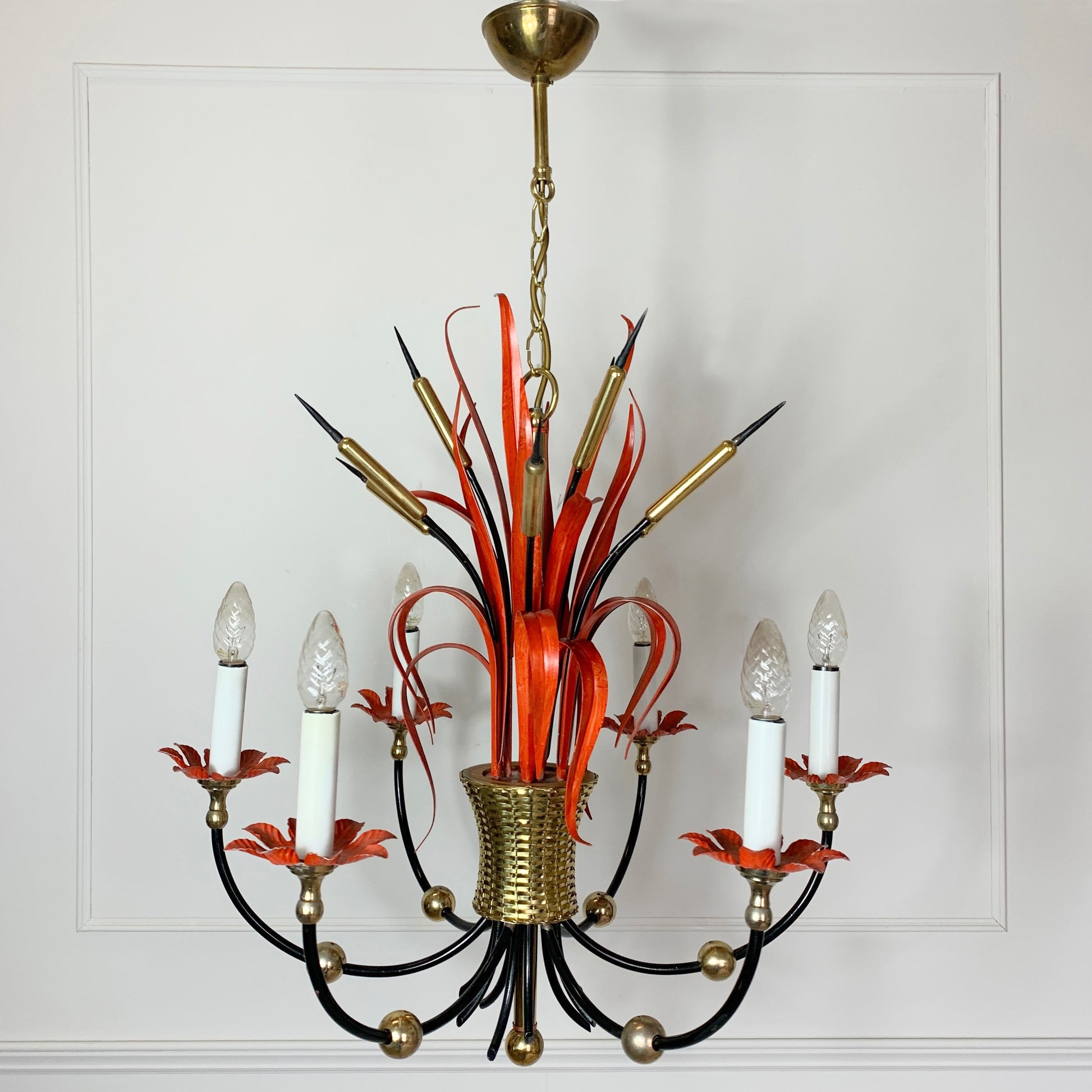 A vintage Maison Bagues Att Red Bulrush Chandelier with a gold-toned brass canopy and central part, black curved arms, orange-red ribbon-like accents, and clear glass candle-style bulbs, shown hanging against a white paneled wall.