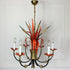 A vintage Maison Bagues Att Red Bulrush Chandelier with a gold-toned brass canopy and central part, black curved arms, orange-red ribbon-like accents, and clear glass candle-style bulbs, shown hanging against a white paneled wall.