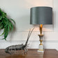 A table lamp with a dark gray fabric drum shade, a gold-toned fluted stem, and a white-and-gray square base, styled on a wooden table beside a lobster figurine and a green fern against a white paneled wall.