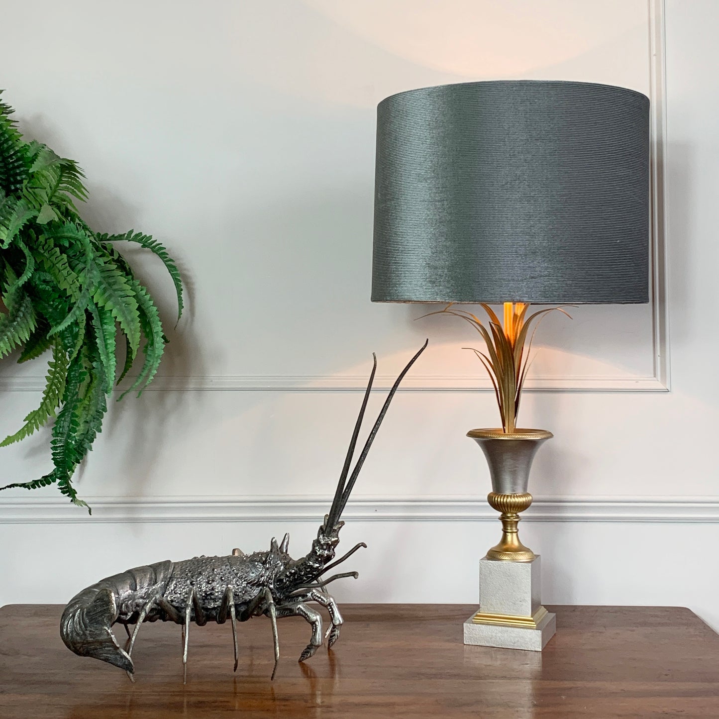 A table lamp with a dark gray fabric drum shade, a gold-toned fluted stem, and a white-and-gray square base, styled on a wooden table beside a lobster figurine and a green fern against a white paneled wall.