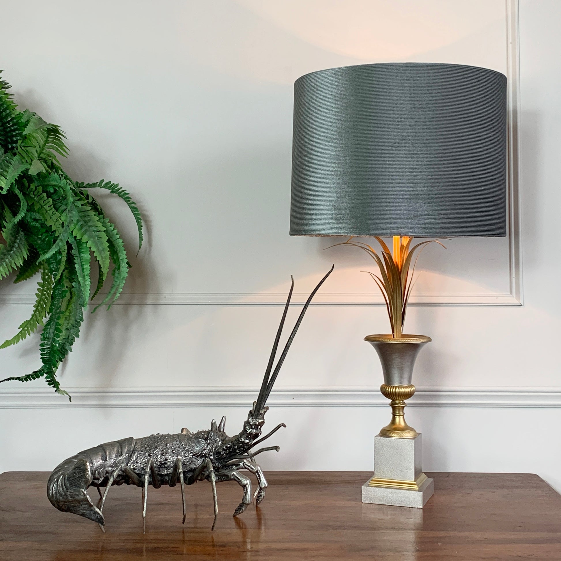 A table lamp with a dark gray fabric drum shade, a gold-toned fluted stem, and a white-and-gray square base, styled on a wooden table beside a lobster figurine and a green fern against a white paneled wall.
