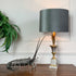 A table lamp with a dark gray fabric drum shade, a gold-toned fluted stem, and a white-and-gray square base, styled on a wooden table beside a lobster figurine and a green fern against a white paneled wall.