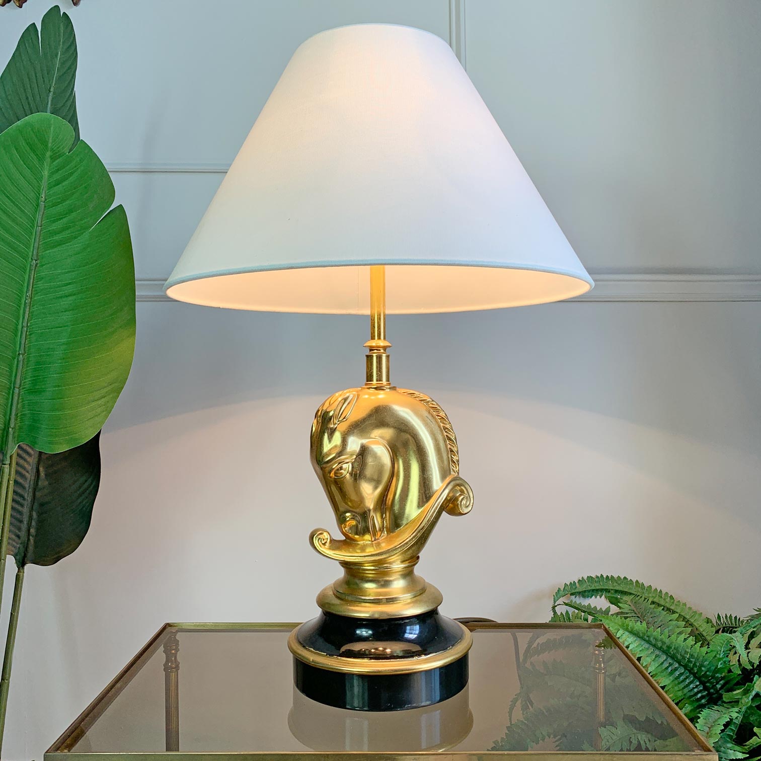 A table lamp with a white conical shade, a polished gold horse-head sculpted base, and a glossy black circular pedestal, shown lit on a glass-topped side table with green plants in the background.