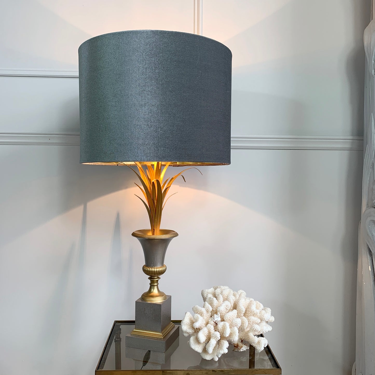 A table lamp with a large textured gray drum shade, a gold-toned fluted stem, and a gray and gold pedestal base, shown lit on a mirrored side table next to a white coral decor piece against a white paneled wall.