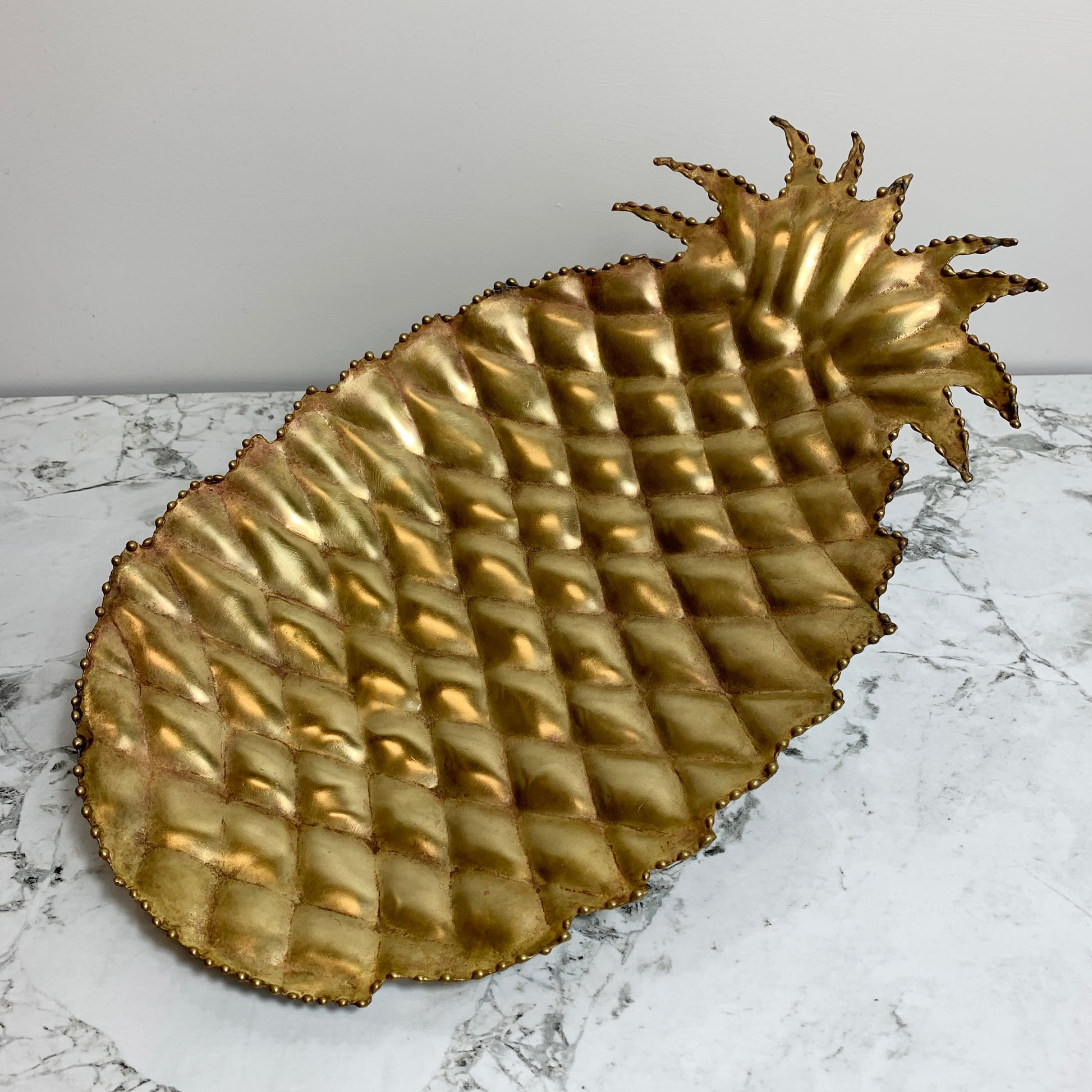 A decorative vintage pineapple-shaped tray or bowl with a metallic gold finish and raised diamond texture, photographed on a white marble surface against a light gray wall.