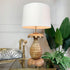 A decorative table lamp with a large white drum shade and a sculptural pineapple-shaped base in a warm natural wood finish, styled on a glass table with plants and a chair in a bright interior setting.