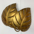 A decorative wall sconce shaped like overlapping gold-toned tropical leaves with cutout details, mounted against a plain white wall.