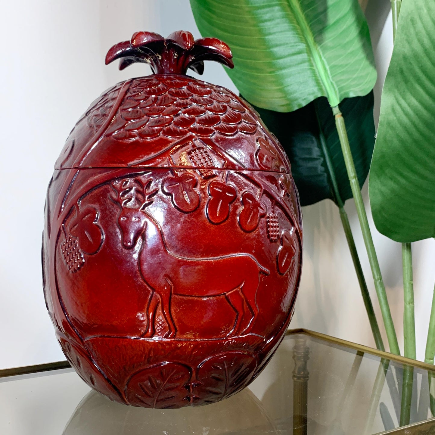 A glossy red Marc Chagall pomegranate-shaped decorative ice bucket with embossed animal and floral details, displayed on a glass tabletop with large green leaves in the background.
