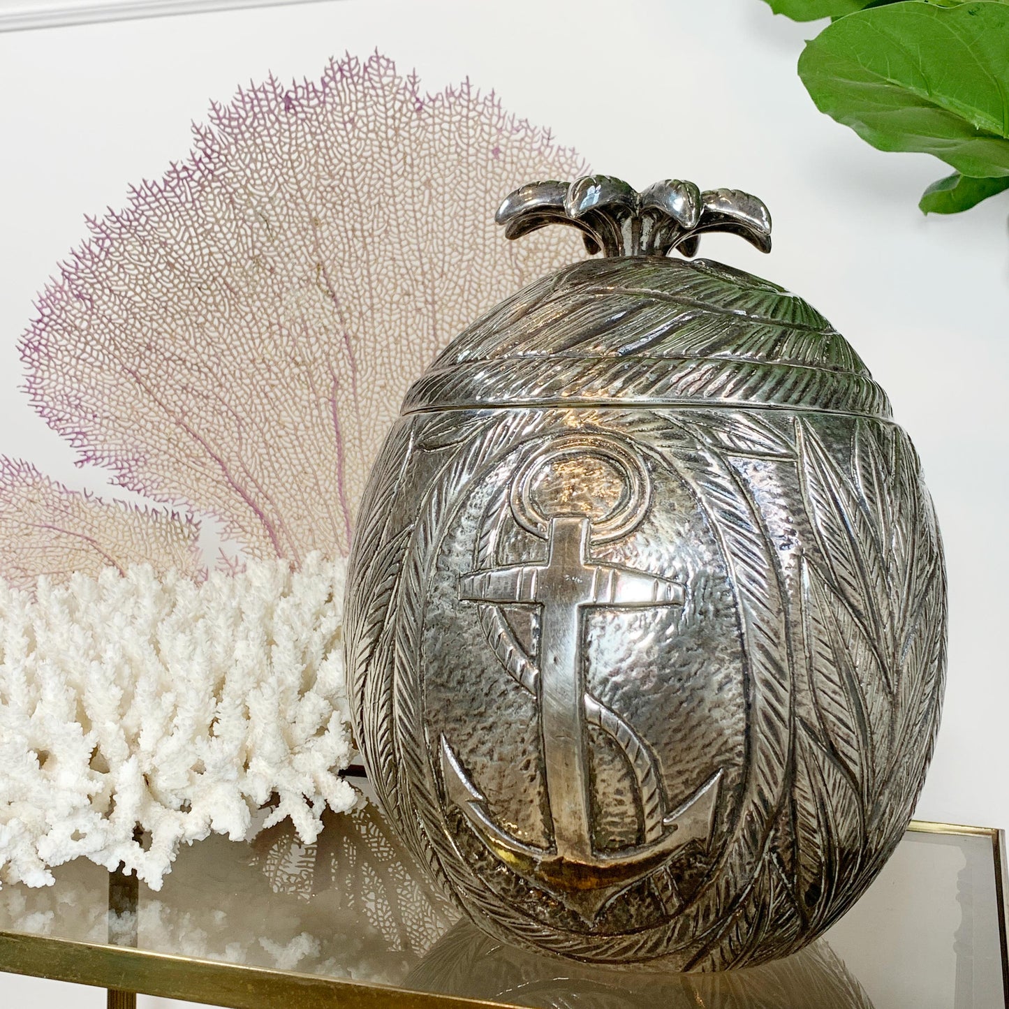 A hammered silver Marc Chagall decorative egg-shaped ice bucket with an embossed anchor and leaf pattern, displayed on a glass shelf with coral and green leaves in the background.