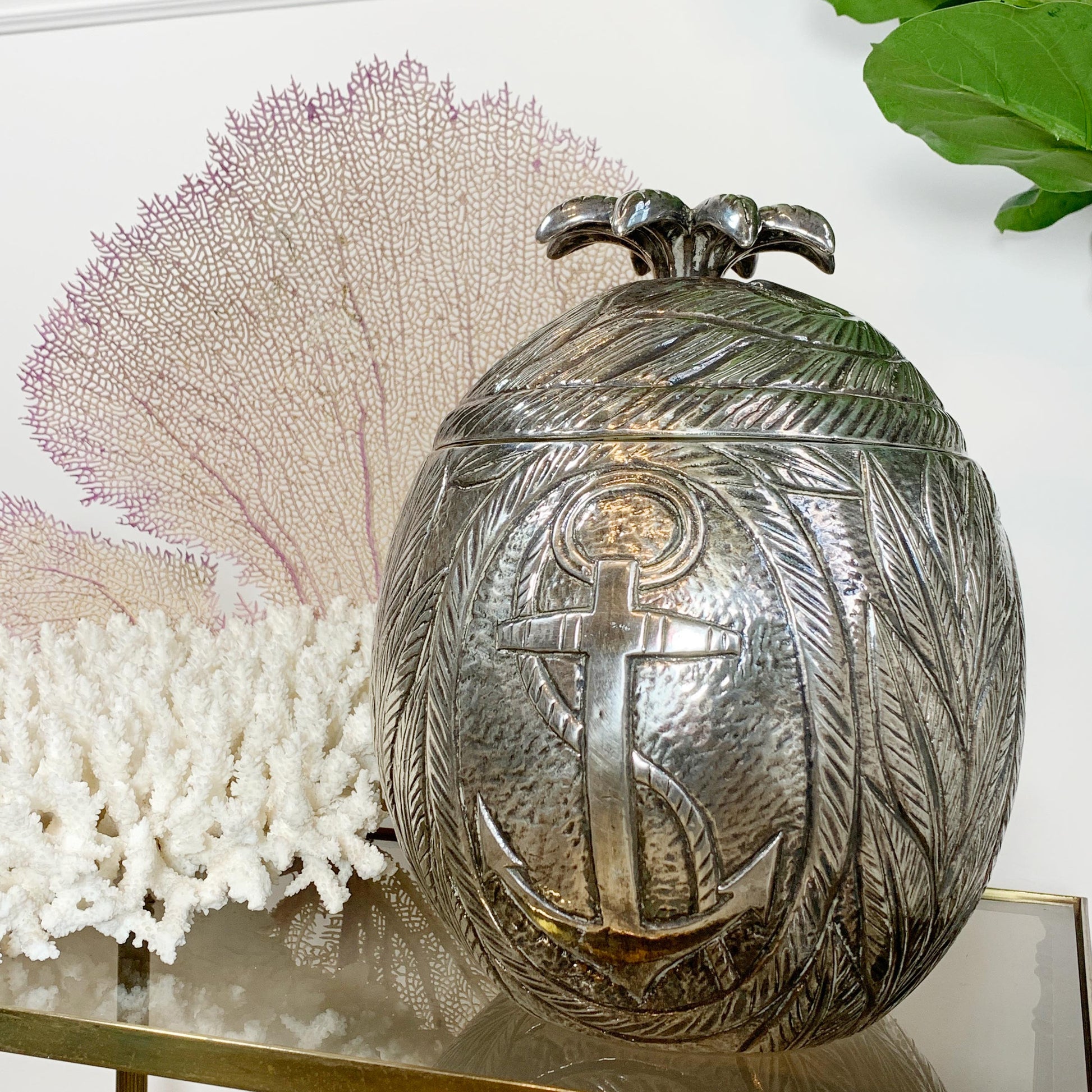 A hammered silver Marc Chagall decorative egg-shaped ice bucket with an embossed anchor and leaf pattern, displayed on a glass shelf with coral and green leaves in the background.