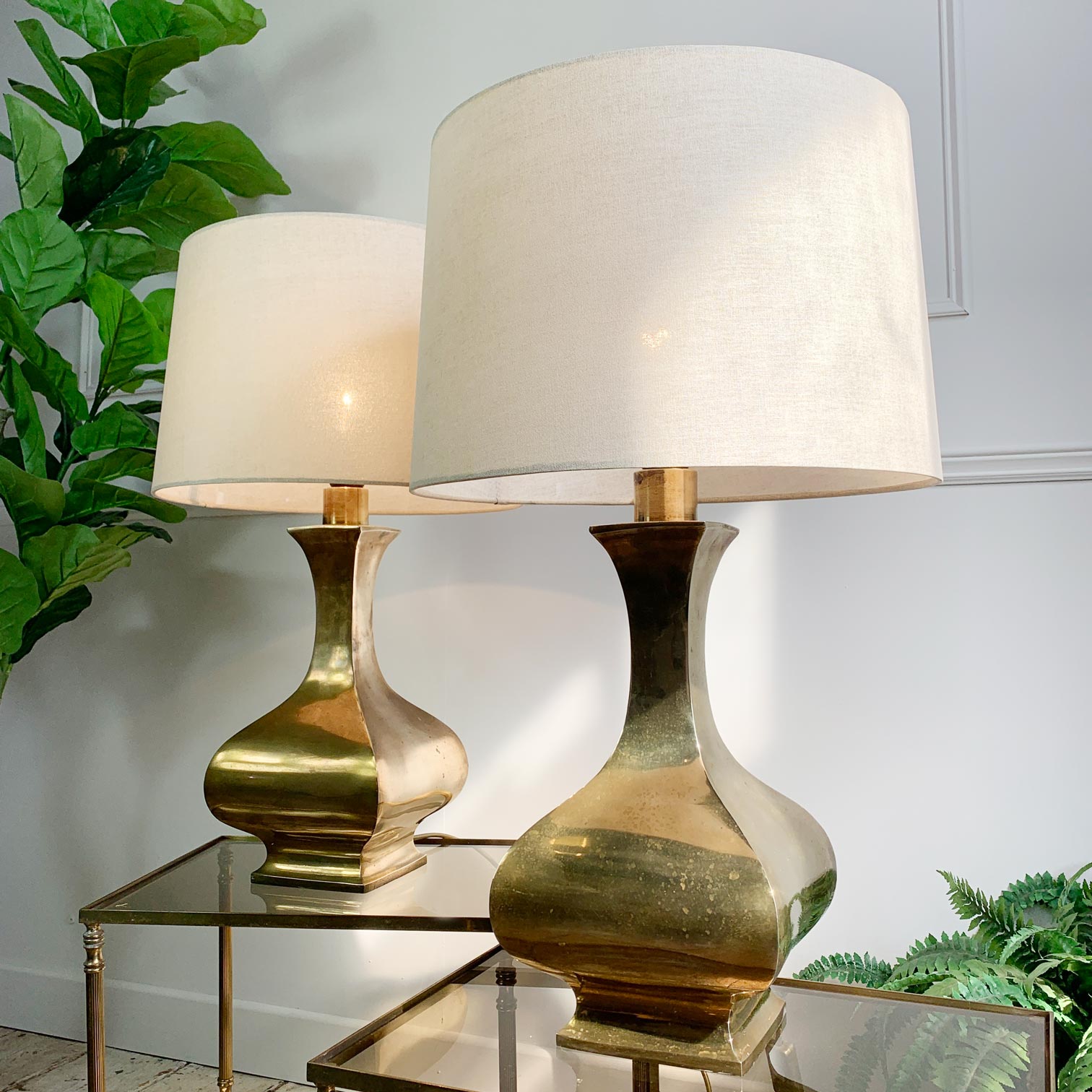 Two table lamps with tapered off-white fabric shades and glossy gold ceramic bases, displayed on a mirrored side table against a white paneled wall with green plants.