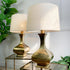 Two table lamps with tapered off-white fabric shades and glossy gold ceramic bases, displayed on a mirrored side table against a white paneled wall with green plants.