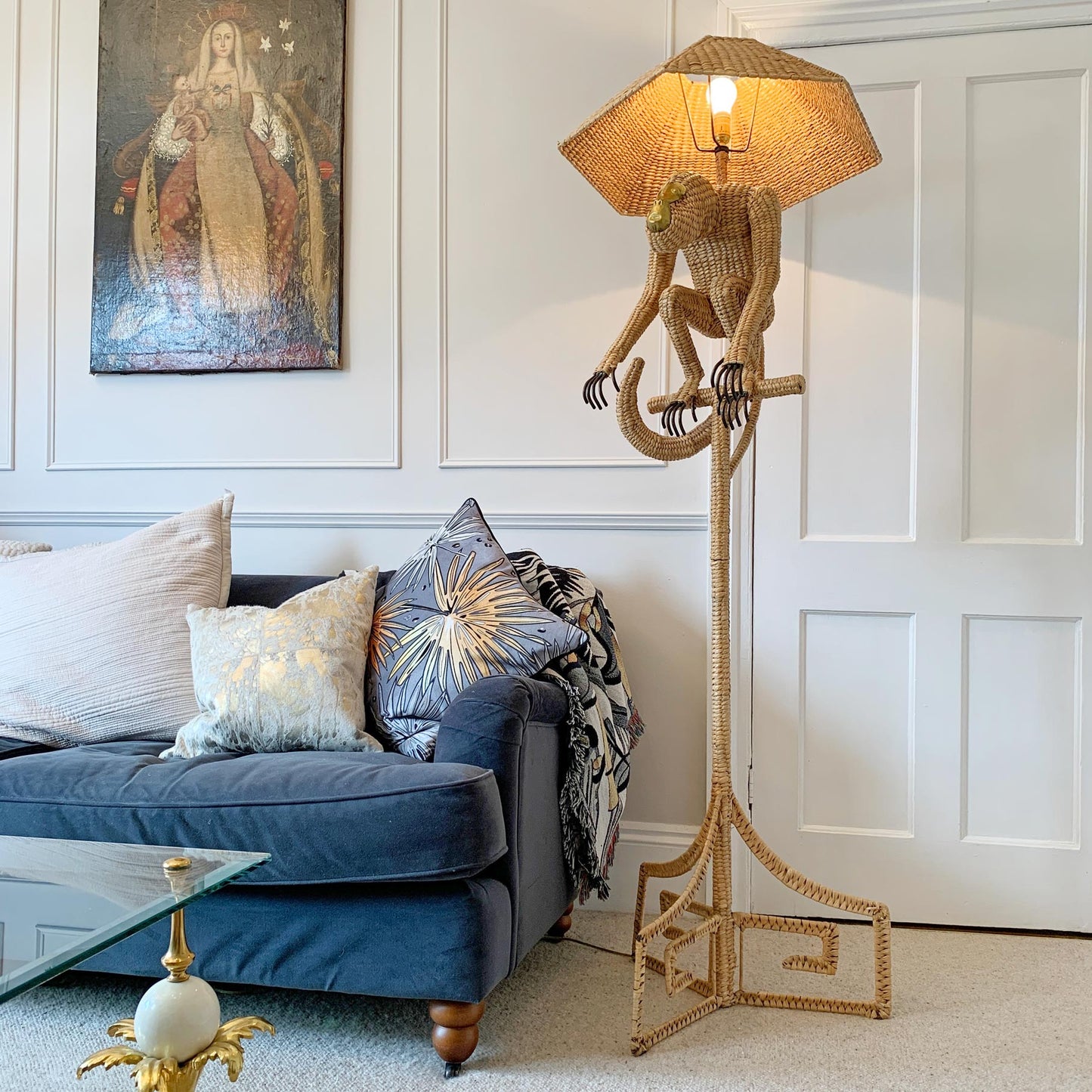 A whimsical floor lamp shaped like a monkey hanging from a coiled rope stand, with a woven wicker umbrella shade, shown lit in a living room next to a blue sofa.