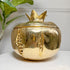 A shiny gold Italian Mauro Manetti pomegranate-shaped decorative ice bucket with a lid, shown on a white surface with a white wall and green plant leaves in the background.