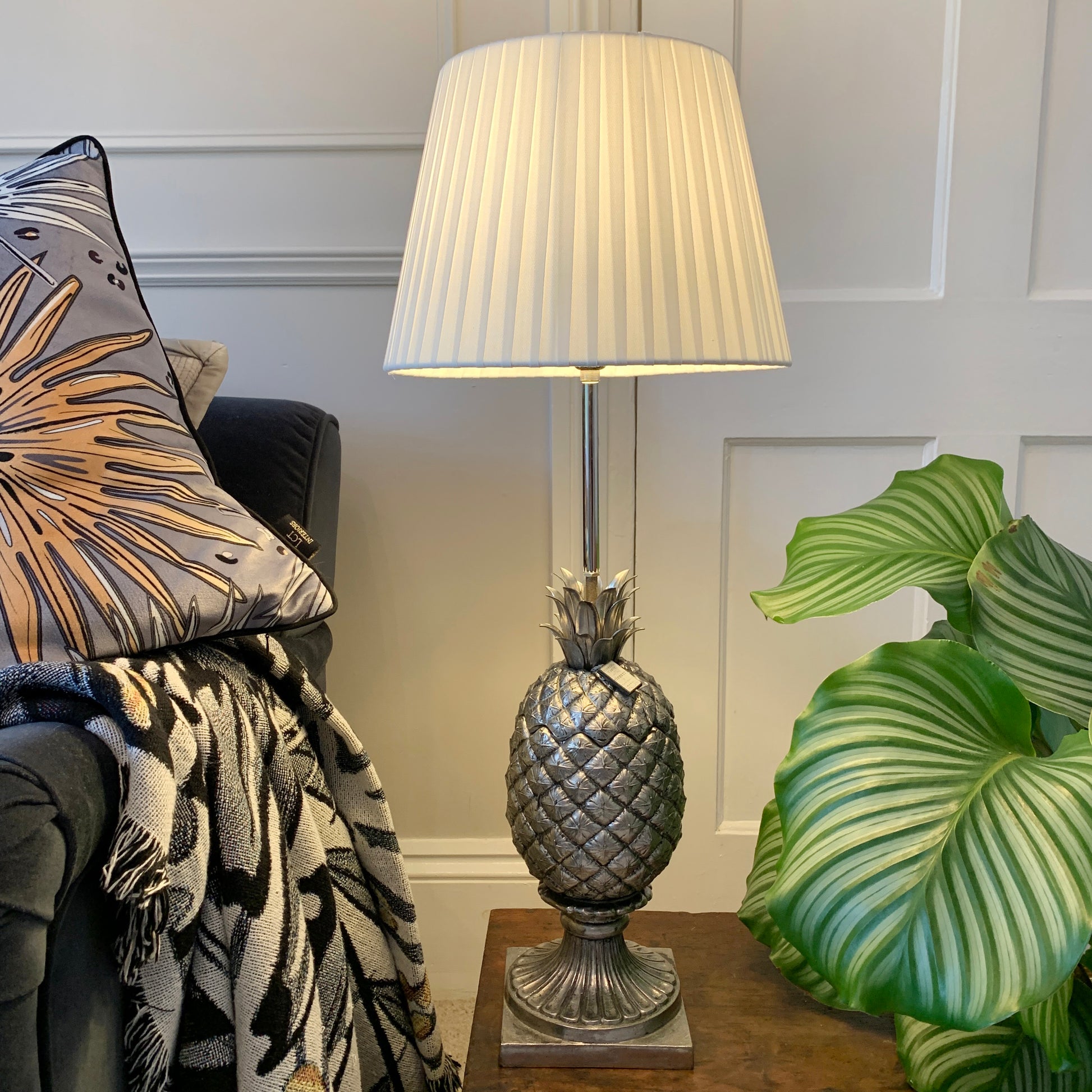 A table lamp with a metallic silver pineapple-shaped base and a pleated off-white fabric shade, styled on a wooden side table next to a green plant and a patterned throw blanket.