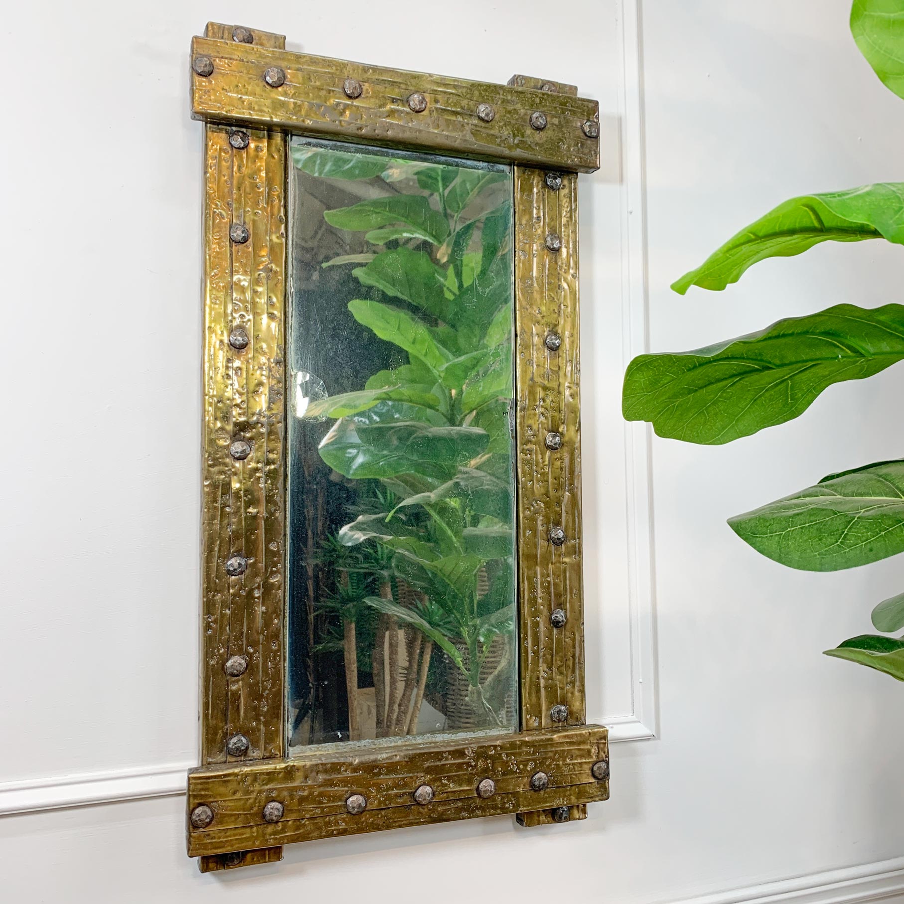 A rectangular brutalist wall mirror with a distressed, rustic bronze-brown frame featuring metal studs, mounted on a white wall next to a green houseplant.