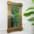 A rectangular brutalist wall mirror with a distressed, rustic bronze-brown frame featuring metal studs, mounted on a white wall next to a green houseplant.