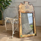 An ornate antique Venetian rectangular mirror with a distressed gold and beige frame featuring floral painted details, leaning against a white wrought-iron chair in an indoor room setting.