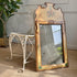 An ornate antique Venetian rectangular mirror with a distressed gold and beige frame featuring floral painted details, leaning against a white wrought-iron chair in an indoor room setting.