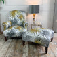 A matching antique upholstered armchair and ottoman set with a white background and LCT Interiors tropical palm leaf fabric in gray and mustard yellow, styled in a room with a wooden floor and a side table with a lamp.