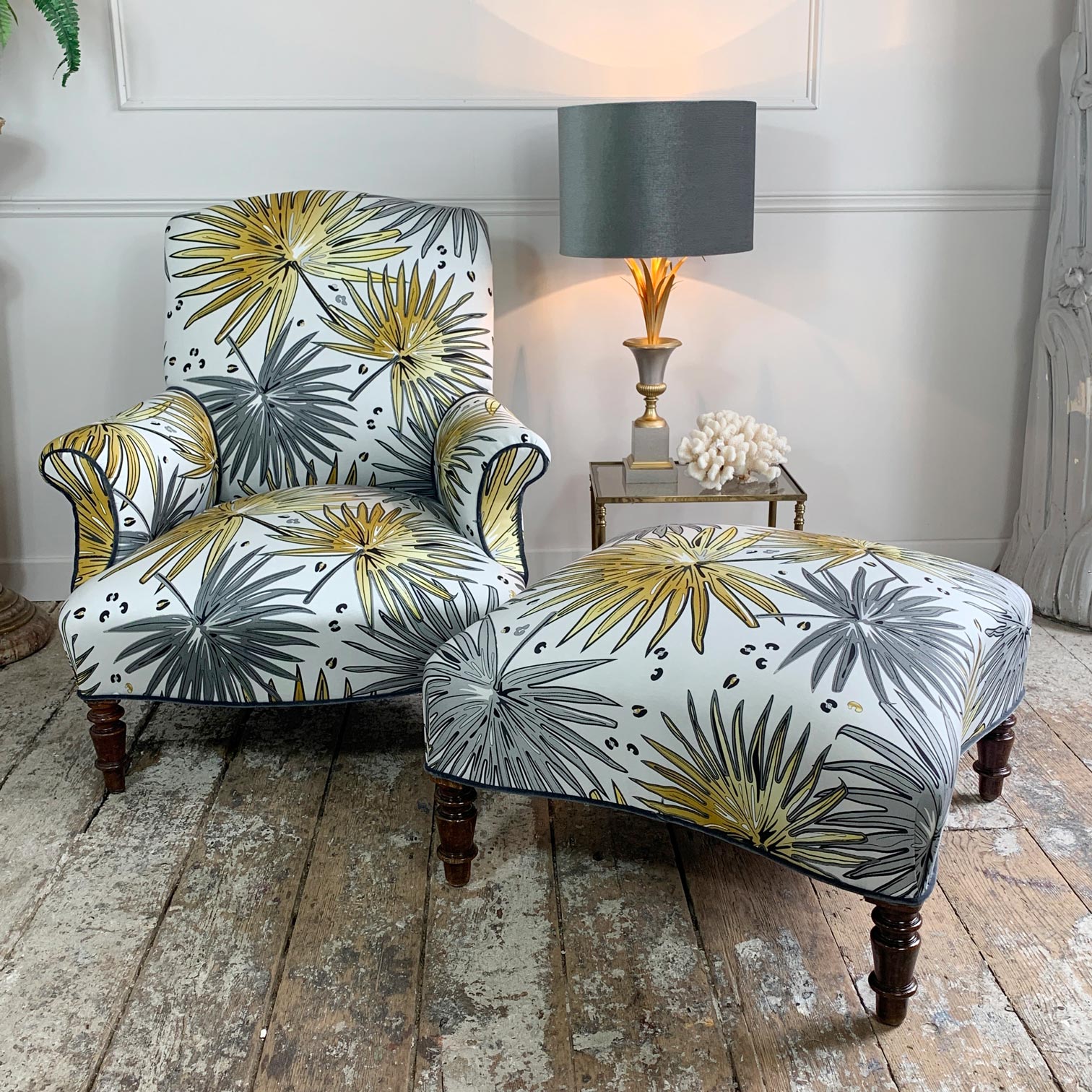 A matching antique upholstered armchair and ottoman set with a white background and LCT Interiors tropical palm leaf fabric in gray and mustard yellow, styled in a room with a wooden floor and a side table with a lamp.