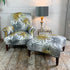 A matching antique upholstered armchair and ottoman set with a white background and LCT Interiors tropical palm leaf fabric in gray and mustard yellow, styled in a room with a wooden floor and a side table with a lamp.