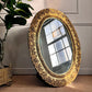 An oval decorative vintage mirror with an ornate carved gold frame, leaning against a paneled wall on a textured rug next to a potted plant.