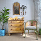 A n antique victorian three-drawer wooden chest of drawers in a warm finish with hand painted details, styled in a room next to a distressed green and beige cane-back chair, a potted fiddle-leaf fig plant, and a decorative wall mirror.