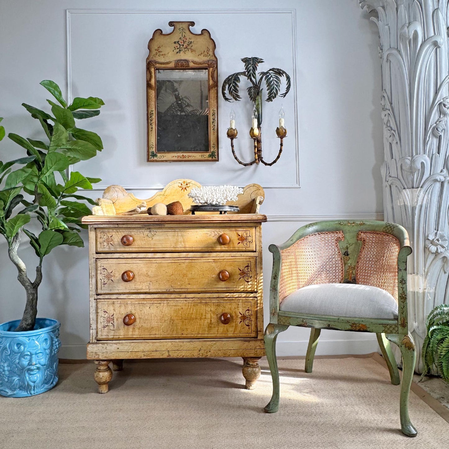 A n antique victorian three-drawer wooden chest of drawers in a warm finish with hand painted details, styled in a room next to a distressed green and beige cane-back chair, a potted fiddle-leaf fig plant, and a decorative wall mirror.