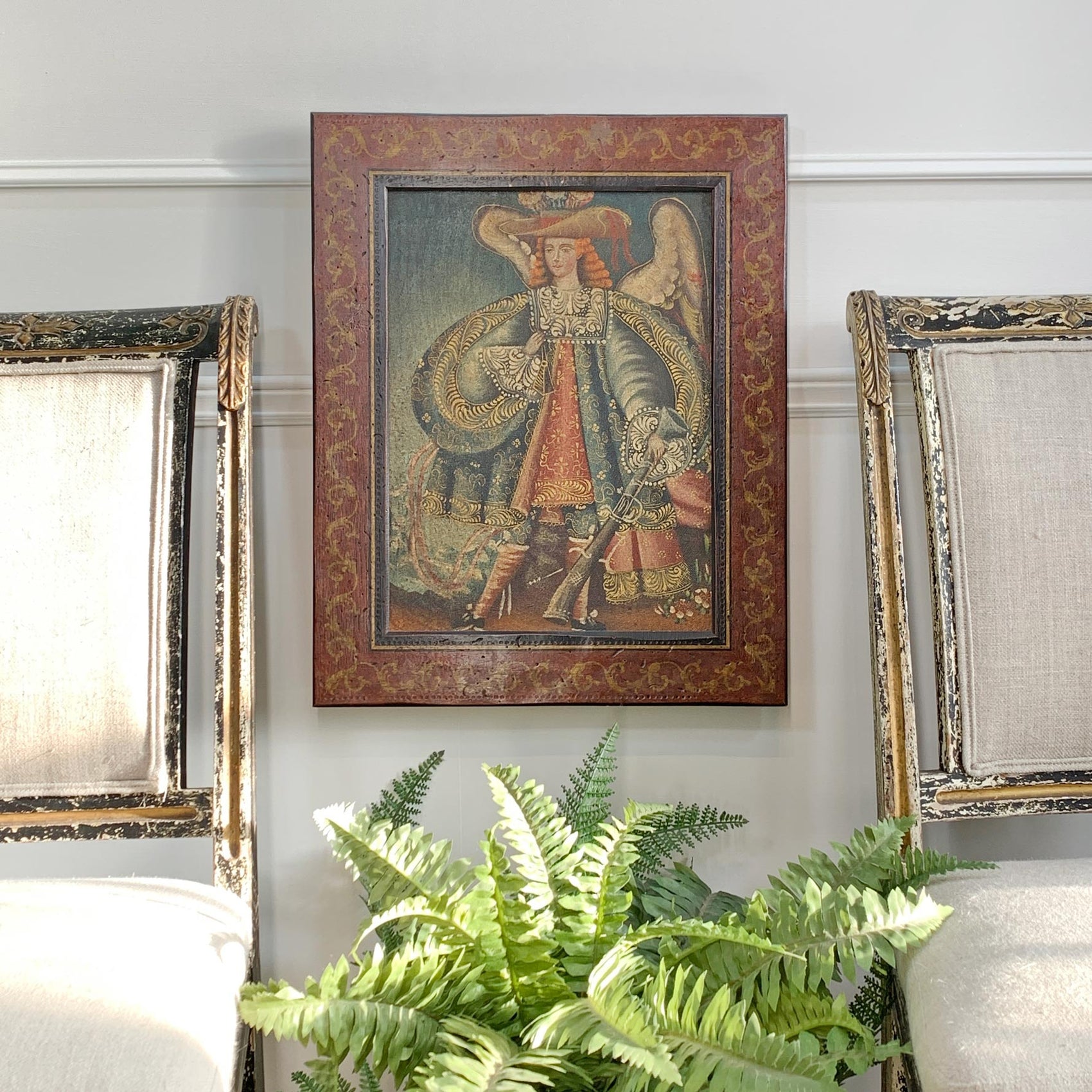 A framed antique portrait painting of Archangel Michael hangs on a light gray wall above a green fern plant, flanked by two ornate framed artworks.