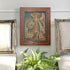 A framed antique portrait painting of Archangel Michael hangs on a light gray wall above a green fern plant, flanked by two ornate framed artworks.