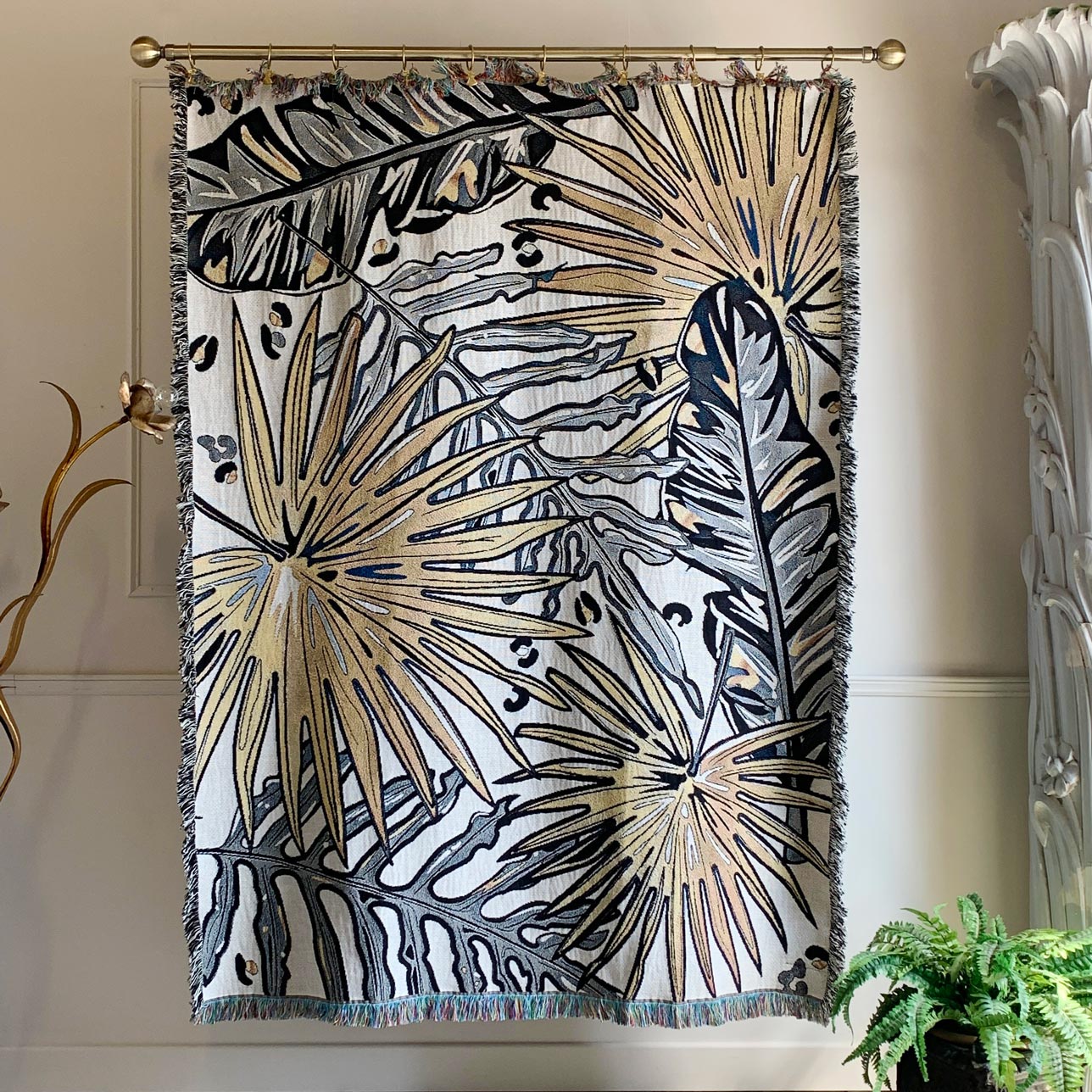 A woven tapestry with a tropical palm leaf pattern in beige, gray, and navy blue, hanging from a gold curtain rod in a home setting.