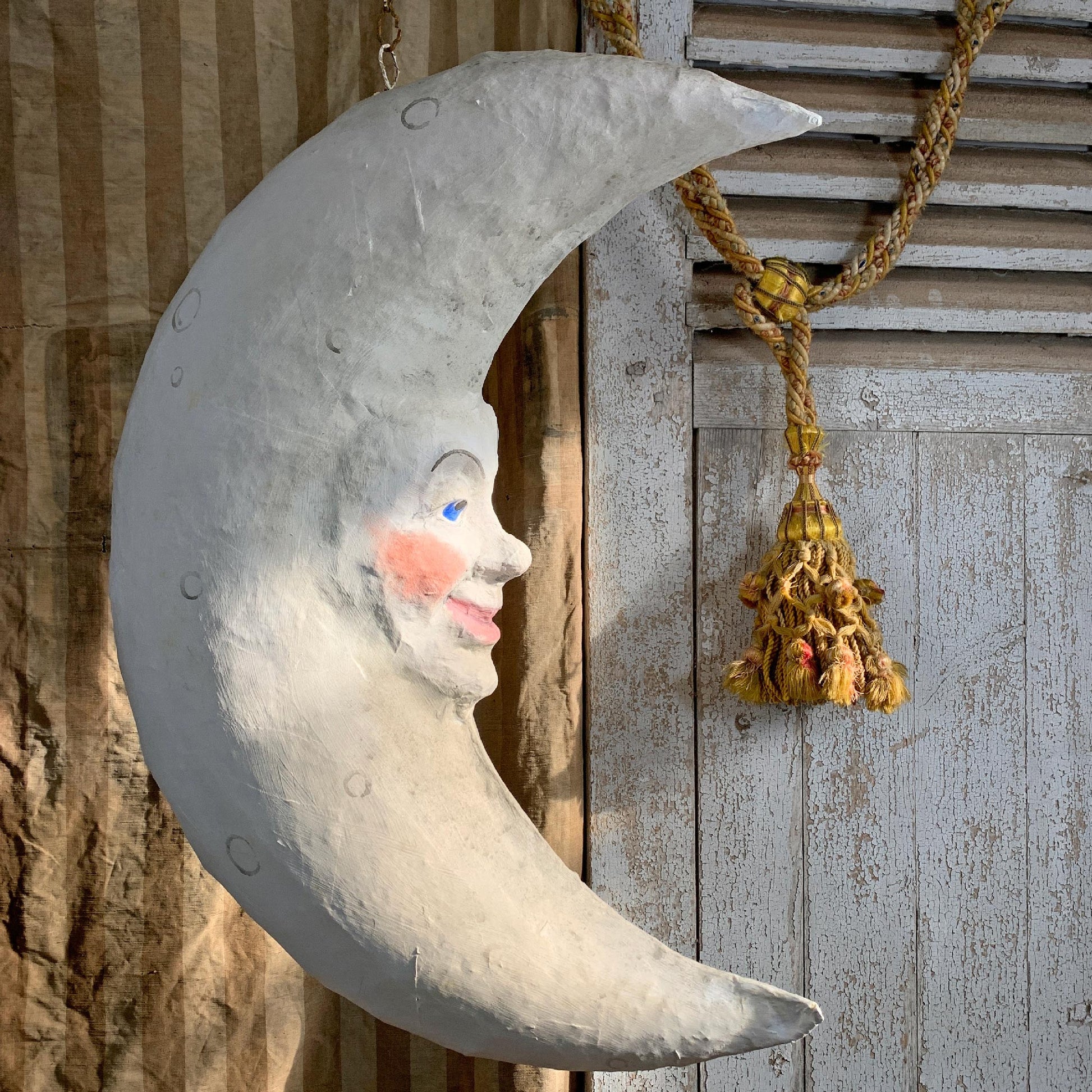 A large paper mache decorative crescent moon wall hanging with a painted moon face, shown hanging against rustic wood panels and a rope with a tassel.