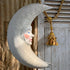 A large paper mache decorative crescent moon wall hanging with a painted moon face, shown hanging against rustic wood panels and a rope with a tassel.