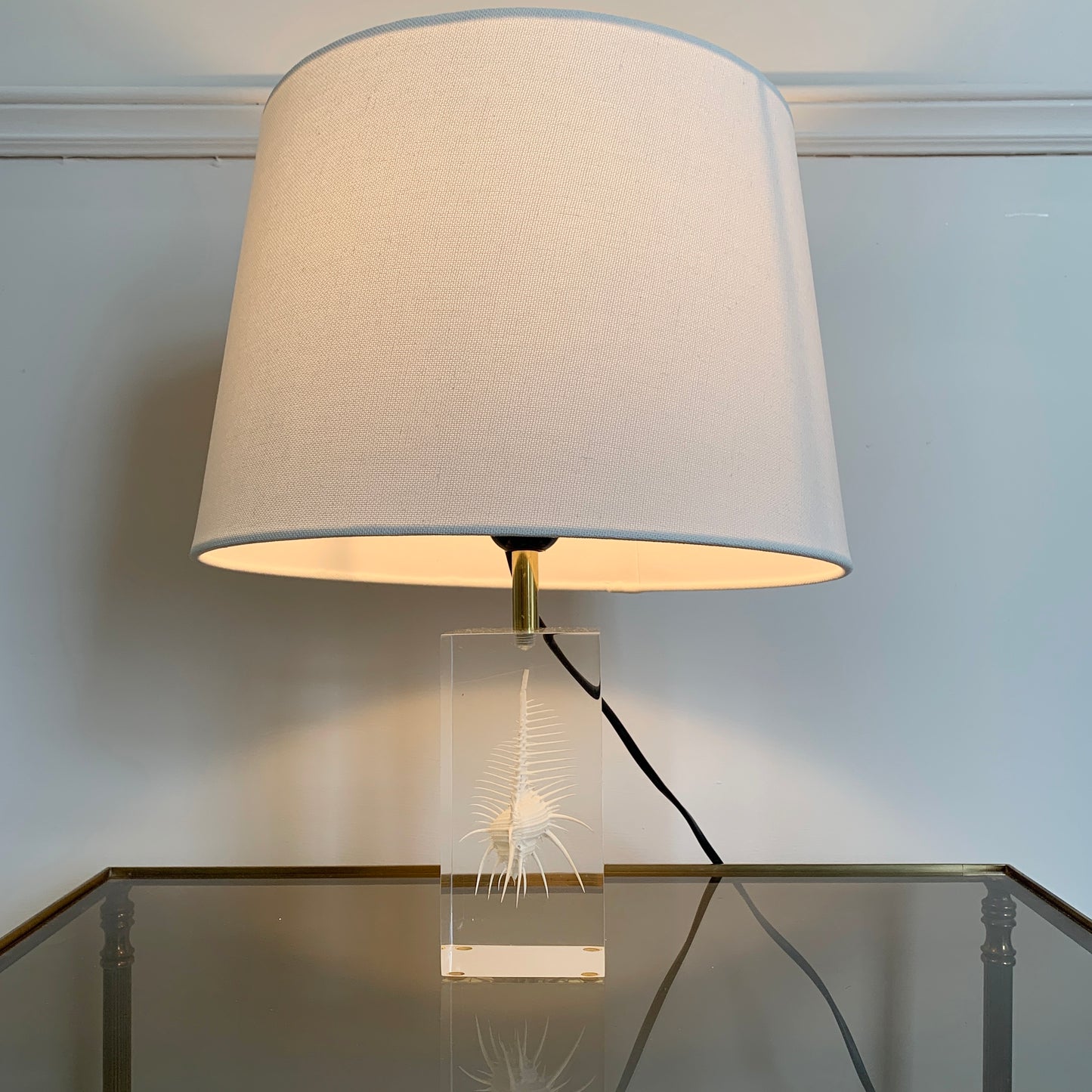 A Pierre Giraudon resin suspension desk lamp comprising a white shell held within a rectangular piece of resin or plexi-glass, and a brass finial, shown lit on a reflective glass table against a white wall.