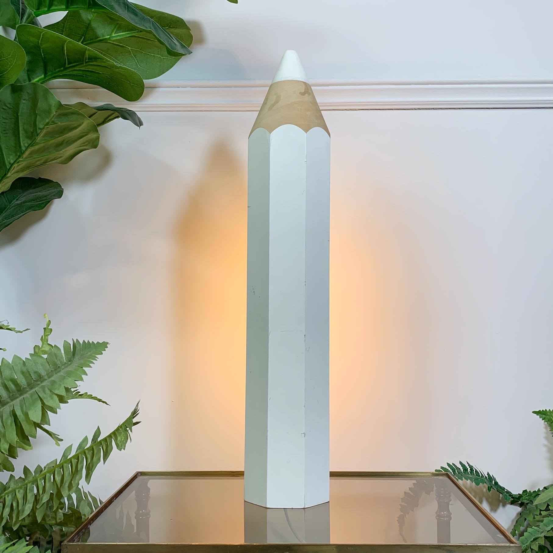 Pierre Sala White Pencil Table Lamp for Vilac 1980's, with a white painted body, a light wood sharpened graphite tip, and a warm glowing bulb inside, displayed on a glass-topped table with green plants in a home setting.