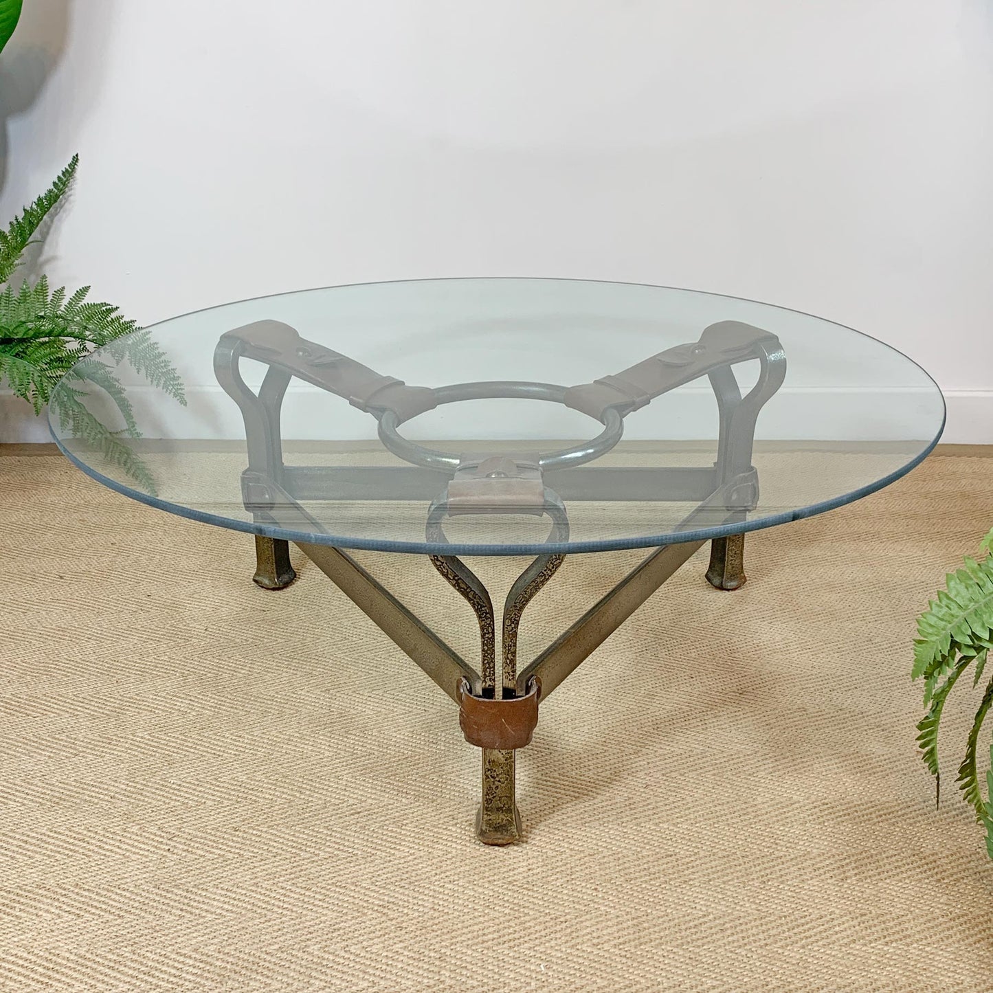Round glass-top designer coffee table with a decorative metal and leather base, shown on a woven beige rug against a white wall with green plants at the sides.