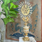 An ornate Processional Gilt Iron IHS Cross Mounted on a Baroque Altar Vase 18th Century with gold and cream detailing, topped with a sunburst and IHS monogram ornament, displayed on a white surface in front of a tapestry and next to a green plant.