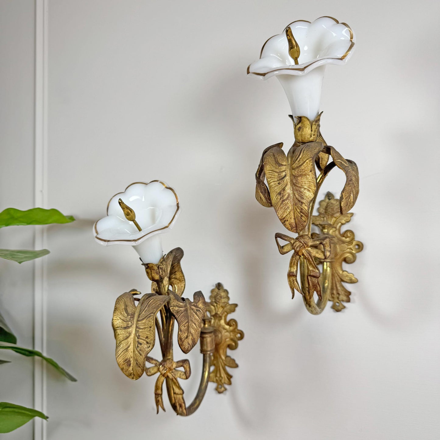 A pair of R W Winfield antique ornate wall sconces with white porcelain calla lily shades and gilded gold leaf accents, mounted on a white wall next to a green plant.