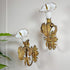A pair of R W Winfield antique ornate wall sconces with white porcelain calla lily shades and gilded gold leaf accents, mounted on a white wall next to a green plant.