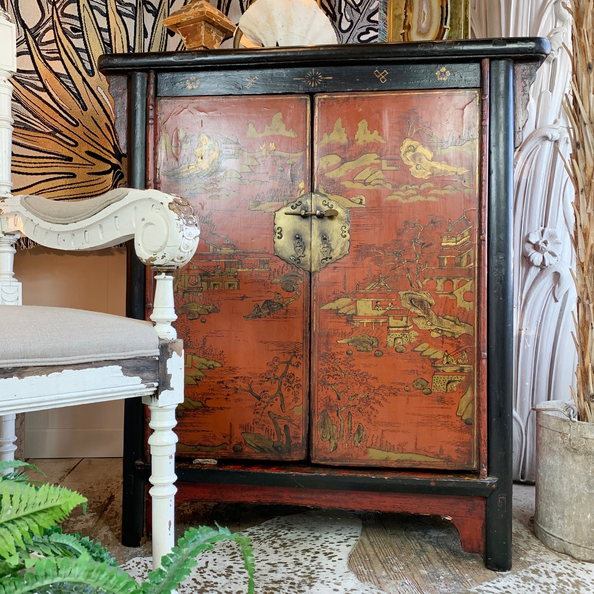 An antique chinoiserie painted cabinet with a reddish-orange lacquered door panel featuring gold scenic illustrations, framed in black wood with ornate brass hardware.