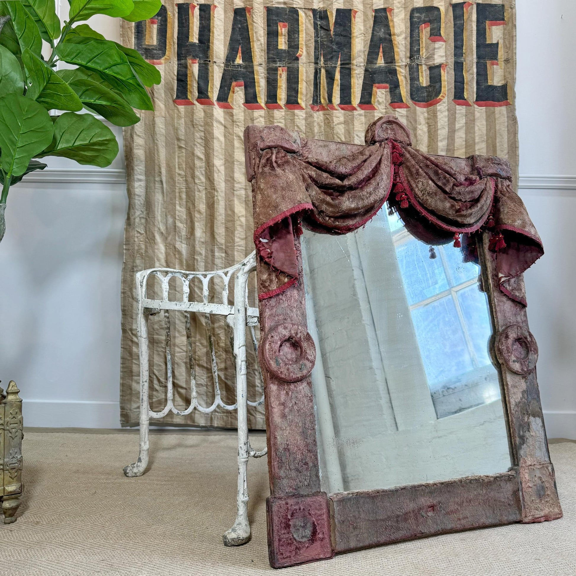 An antique rectangular mirror with a swag velvet, weathered reddish-brown frame and a draped fabric valance across the top, leaning against a beige wallpapered wall next to a white metal stand.