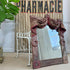 An antique rectangular mirror with a swag velvet, weathered reddish-brown frame and a draped fabric valance across the top, leaning against a beige wallpapered wall next to a white metal stand.