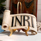 A decorative scroll-shaped wooden religious church carving with the letters 'INRI' in raised black font on an off-white distressed background, displayed on a gold stand.