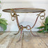 An oval metal designer end table with a distressed gold and reddish-brown painted frame, set on a rustic wooden floor against a white wall with green fern plants on either side.