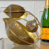 A decorative gold-toned metal and glass leaf-shaped serving sculpture with a textured chestnut center piece, displayed on a reflective surface next to a champagne bottle against a white paneled wall.
